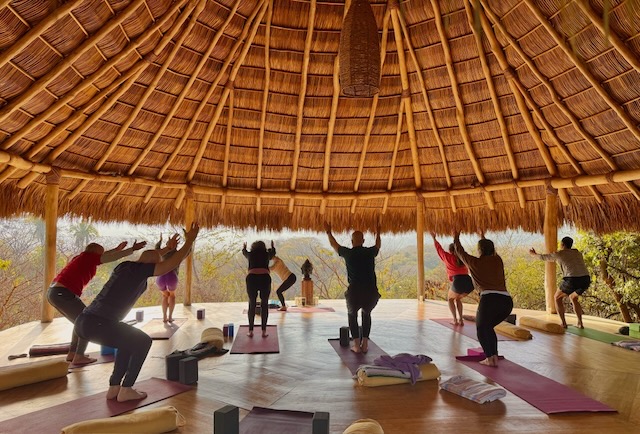 Group practicing yoga under a large straw hut at the Rooted Connection Couples Retreat